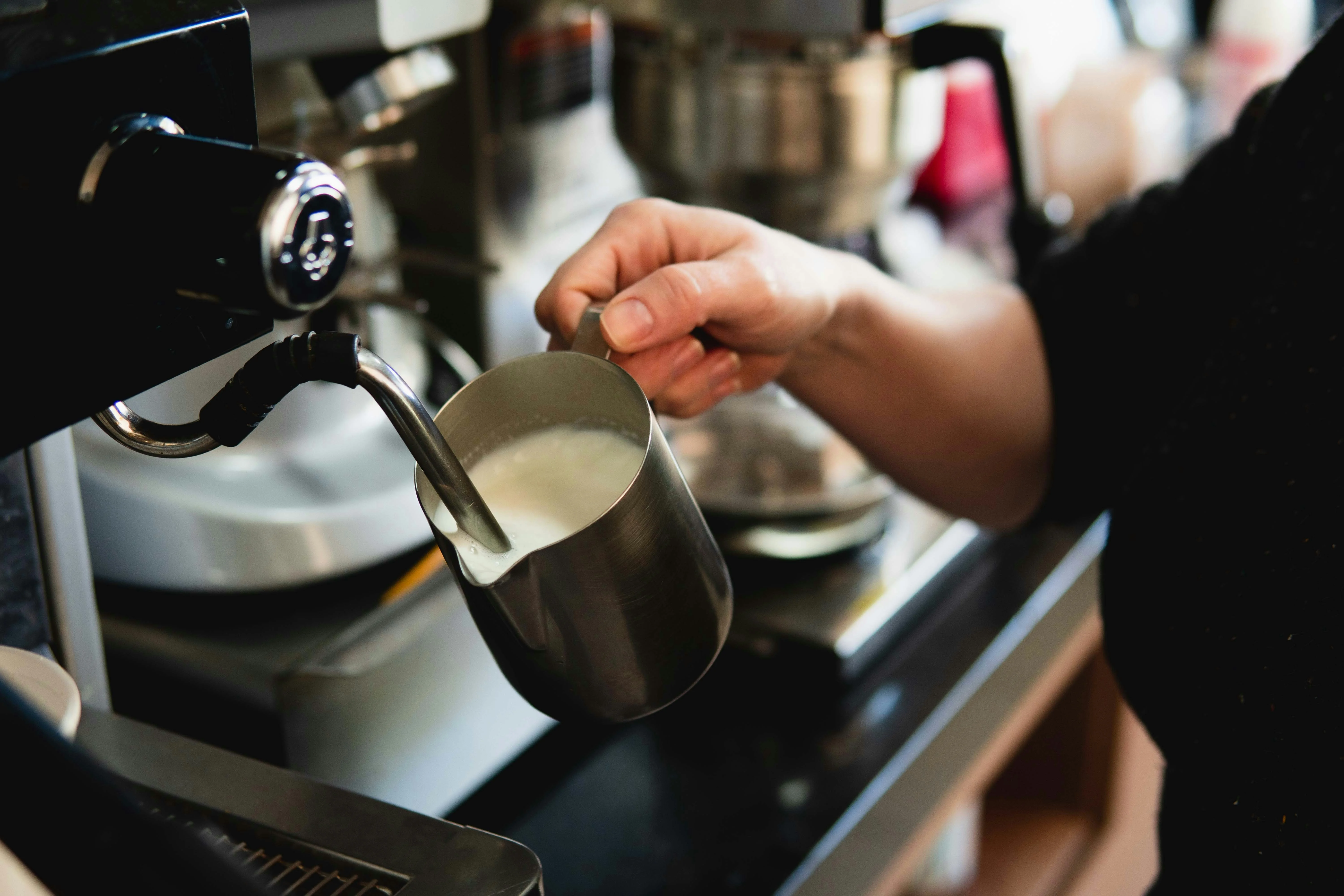 Close-up of a steam wand positioned just below the surface of milk in a pitcher, angled to create a whirlpool